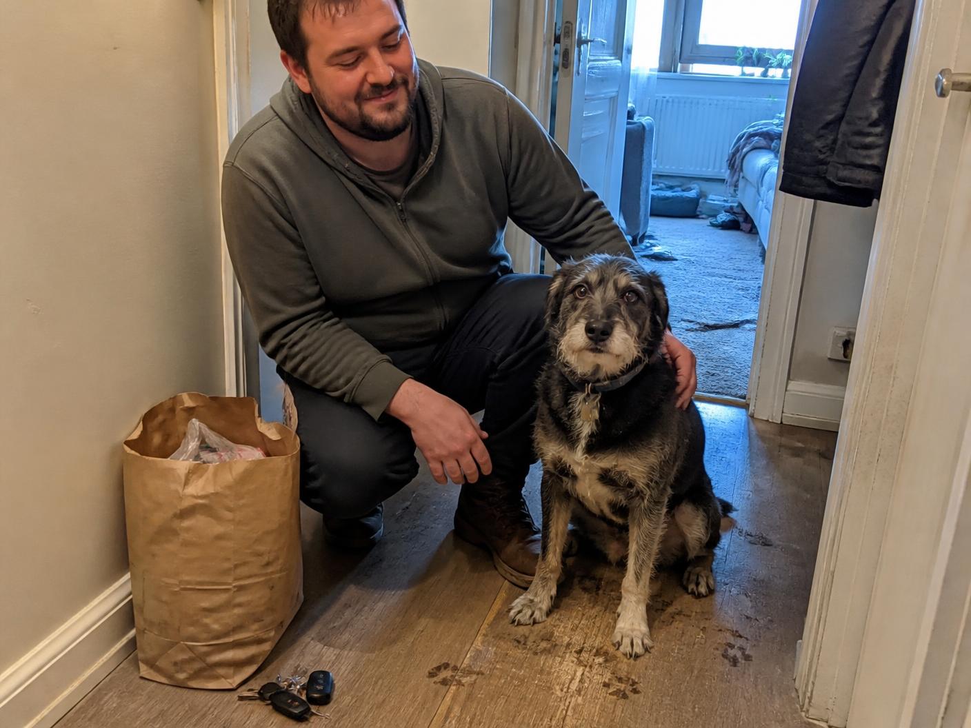 A man kneeling beside his scruffy mixed-breed dog in an everyday apartment hallway